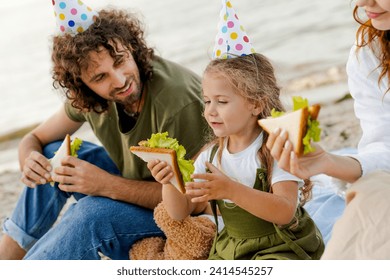 Close up shot of family with party hats eating sandwiches on the beach. Family enjoying daughter's birthday with snacks outdoors. Healthy eating, vegetarian meal. Food festival - Powered by Shutterstock