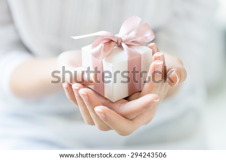 Close up shot of female hands holding a small gift wrapped with pink ribbon. Small gift in the hands of a woman indoor. Shallow depth of field with focus on the little box.