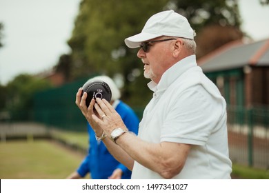 A Close Up Shot Of A Focused Senior Man Holding A Bocce Ball, Ready To Take His Shot. He Is Wearing A White Cap And Sunglasses.