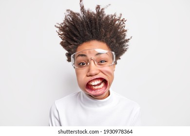 Close up shot of funny young Afro American woman with curly hair standing up has wide opened mouth wears transparent glasses casual turtleneck poses indoor exclaims loudly. Flight of imagination