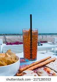 Close up shot of a glass of Spritz, italian aperitif, and complimentary snack, potato crisps, pizza, grissini and salami slices. Beach and sea in the background, Riviera Romagnola, Adriatic Sea.