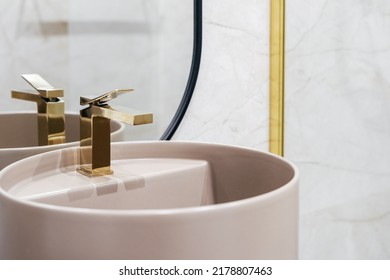 Close up shot of golden shiny water tap over washbasin. Pink wash basin with faucet in classic style in light bathroom against copy space on tiled wall. Concept s of morning routine, hygiene at home