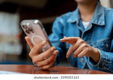 The close up shot of a hand in denim jacket holding a smartphone with left hand and pointing at the screen with right hand. sitting on wooden table.