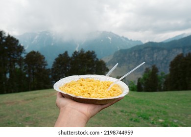 Close up shot of hand holding dish of noodles in background of mountains in Manali, Himachal Pradesh, India. Tourist enjoying Food in mountains during holiday vacation.  