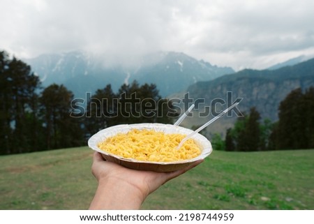 Close up shot of hand holding dish of noodles in background of mountains in Manali, Himachal Pradesh, India. Tourist enjoying Food in mountains during holiday vacation.  