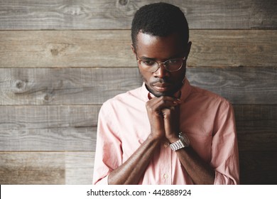 Close up shot of handsome young African man praying and meditating against wooden background. Dark skinned student wearing pink shirt, holding hands in prayer against his lips, hoping for the best