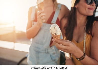 Close up shot of ice cream in hand of a woman standing with her friend. Two young women outdoors eating icecream on a sunny day.