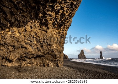 Close up shot from inside a volcanic cave featuring a spectacular jagged basalt ceiling. In the background, the Reynisfjara black sand beach opens up with the rough ocean waves and Reynisdrangar sea stacks under a clear blue sky.