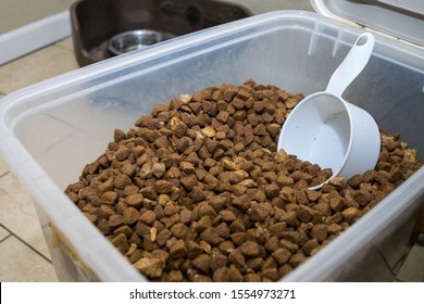 Close Up Shot Of A Large Plastic Container Filled With Dog Food, Kibble And Freeze Dried Meats. Scooper In Food, Water Bowls In Background.