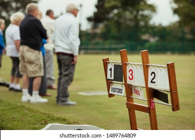 A close up shot of a lawn bowling scoreboard on grass, with senior adults spectating the lawn bowling game.