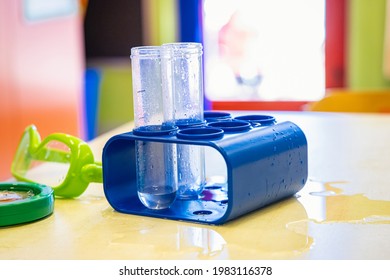 Close Up Shot Of A Little, Plastic Toy Lab Ampoule Filled With Water, On The Colorful Table Of An Indoor Play Area In A Kindergarten.