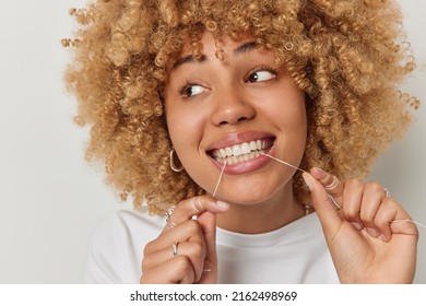 Close Up Shot Of Lovely European Woman Uses Dental Floss Cleans Teeth Undergoes Hygiene Routines Dressed In Casual T Shirt Focused Away Isolated Over White Background. Teeth Flossing Concept