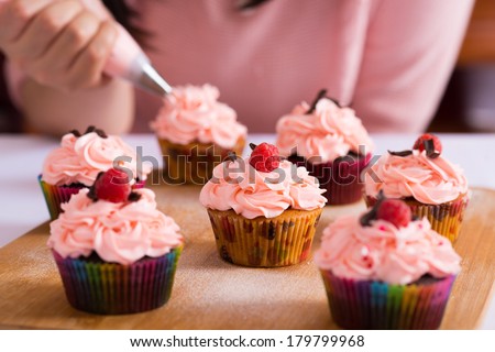 Close shot of many sweet cupcakes on the foreground while a baker decorating the last one 