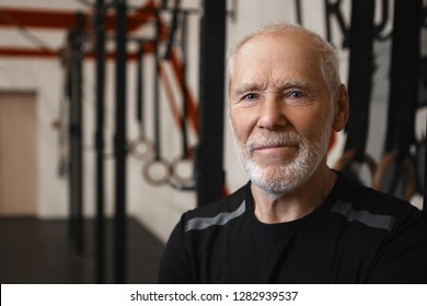 Close Up Shot Of Mature Seventy Year Old Caucasina Man With Wrinkles, Blue Eyes And Thick Beard Wearing Stylish Black T-shirt While Exercising In Gym Alone, Looking At Camera With Happy Smile