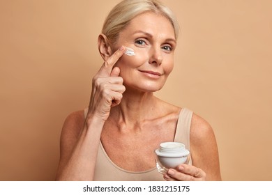 Close up shot of middle aged beautiful woman applies anti aging cream on face undergoes beauty treatments cares about skin poses against beige background. Wrinkled female model with cosmetic product - Powered by Shutterstock