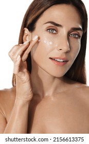 Close Up Shot Of Middle Aged Beautiful Woman Applies Anti Aging Cream On Face Undergoes Beauty Treatments Cares About Skin Poses Against Beige Background. Wrinkled Female Model With Cosmetic Product