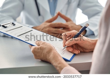 Close up shot of older senior female patient hands holding pen filling medical heath data form sitting at desk with family doctor in modern hospital clinic. Healthcare illness prevention concept.