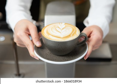 Close up shot on front view of barista girl hands serving beautiful flower shape on fresh latte art in coffee cup on dark grey plate pass over coffee maker machine at coffee shop and cafe restaurant. 