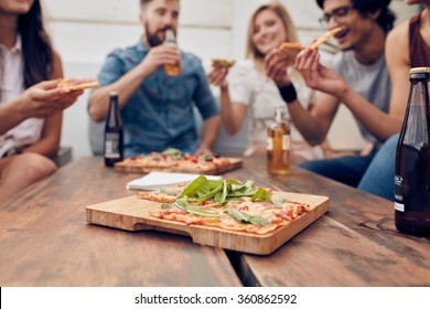 Close up shot of pizza on wooden plate with people eating and drinking in background. Group of friends gathered around the table at a party.