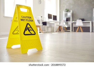 Close Up Shot Of Plastic Standing Sign That Reads Caution Wet Floor And Has Figure Of Man Who Slips And Falls Office Interior In Blurred Background. Commercial Janitorial And Cleaning Service Concept