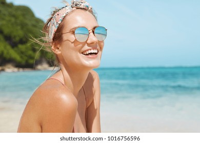 Close up shot of pleasant looking female wears headband and sunglasses, travels in tropical country, poses against warm peaceful ocean, blue sky and rocky cliff in background. Good relaxation - Powered by Shutterstock