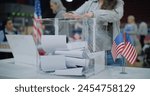 Close up shot of polling box standing on table in polling station. American female voter with kid on hands puts bulletin into box. Political races of US presidential candidates. National Election Day.