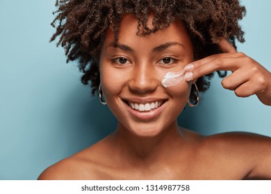 Close Up Shot Of Positive Dark Skinned Woman With Curly Hair, Appiles Face Cream On Cheek, Poses Nude Against Blue Background, Cares Of Beauty, Uses Cosmetic Product. Natural Beauty Concept.