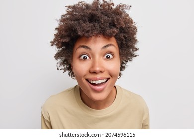 Close up shot of pretty cheerful woman gazes with happy surprised expression at camera smiles toothily wears casual t shirt isolated over white background looks with big interest at somethning amazing
