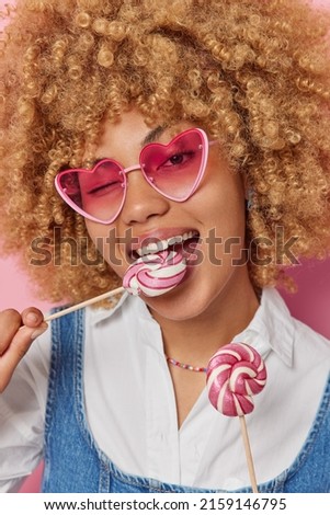 Close up shot of pretty curly haired woman bites caramel candy has sweet tooth wears tredny heart shaped sunglasses and fashionable outfit eats lollipops. Unhealthy eating and harmful food concept