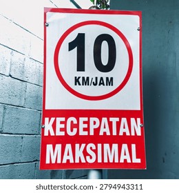 A close up shot of a rectangular Indonesian traffic sign showing a maximum speed limit of ten kilometers per hour with the words kecepatan maksimal posted outside on a pole between a white brick wall and a teal door