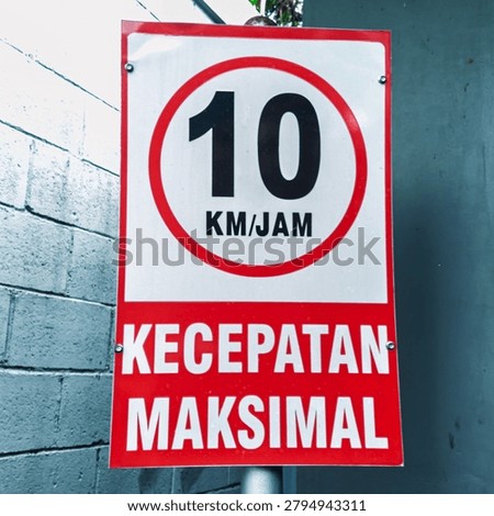A close up shot of a rectangular Indonesian traffic sign showing a maximum speed limit of ten kilometers per hour with the words kecepatan maksimal posted outside on a pole between a white brick wall and a teal door