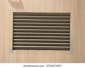 A close up shot of a rectangular ventilation grille with horizontal louvers installed on a light-colored wooden or laminate wall panel
