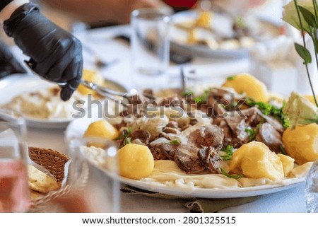 Close up shot of a restaurant server in black protective glove using tongs to portion authentic Central Asian Beshbarmak with boiled meat, horsemeat sausage kazy, tender dough sheets, and potatoes