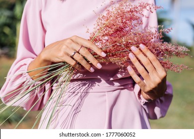 Close shot of romantic cute pink gel polished manicure nails. Woman in pink summer dress holding wild flowers. Two rings on fingers. Outdoor portrait.
