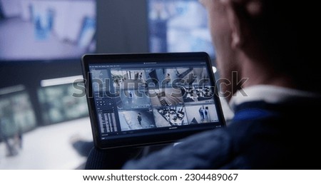 Close up shot of security officer zooming CCTV cameras with AI facial scanning using tablet in police surveillance center. Colleagues work at background. Monitoring system. Concept of social safety.