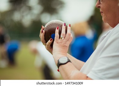 A close up shot of a senior woman holding a bocce ball, ready to take her shot. She is wearing nail polish and a watch.