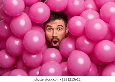 Close up shot of shocked bearded European man looks with widely opened mouth at camera being greatly surprised poses over inflated pink balloons impressed to get unexpected gift on birthday.