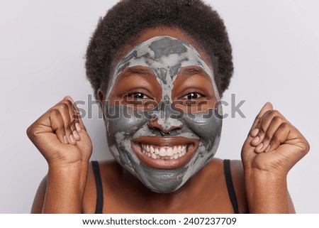 Close up shot of short haired woman with dark skin applies facial clay mask for skin rejuvenation clenches fists and smiles broadly feels very happy isolated over white background. Beauty procedures Foto d'archivio © 