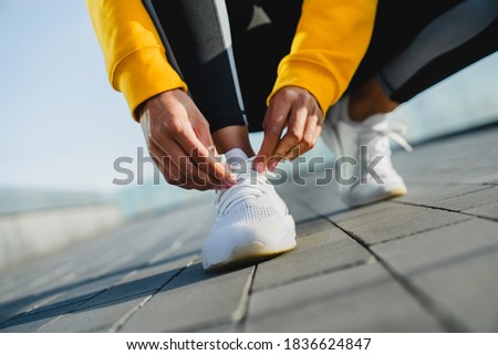 Close up shot of a slim female jogger sitting and tying her shoelaces on white sneakers outside
