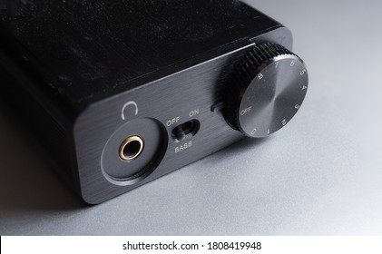 Close Up Shot Of A Small Digital Audio Converter, Or DAC, On A Silver Metallic Surface.  Volume Dial, Headphone Jack, And Bass Boost Switch.  Black Metal Device For Audiophiles.