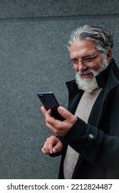 Close Shot Of A Smiling And Confident Mature Businessman Walking Outside While Going To A Meeting And Using A Smartphone. Senior Bearded Man Dressed In Stylish Black Coat Holding Mobile Phone.