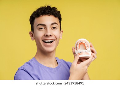 Close up shot of a smiling, curly haired teenager with braces, holding dental molds, laughing, isolated on yellow background. Emphasizes orthodontic therapy concept  - Powered by Shutterstock