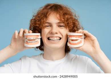 Close up shot of a smiling, curly red haired teenager with braces, playfully holding dental molds, laughing, isolated on a blue background. Emphasizes orthodontic therapy concept - Powered by Shutterstock