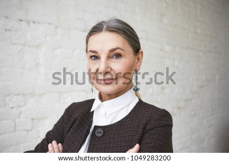 Close up shot of successful beautiful confident senior businesswoman in her fifties with gray hair and blue wise eyes posing indoors, keeping arms folded, looking at camera with charming smile