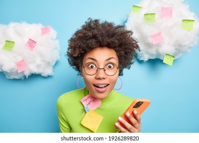 Close Up Shot Of Surprised Curly Woman Stares Surprisingly At Camera Holds Mobile Phone Checks Email Box Surrounded With Memo Notes Makes List To Do Tries To Be More Productive Poses Indoor.