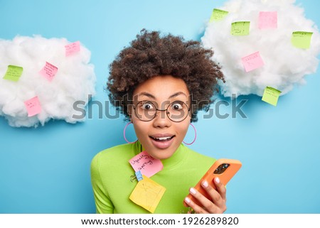 Close up shot of surprised curly woman stares surprisingly at camera holds mobile phone checks email box surrounded with memo notes makes list to do tries to be more productive poses indoor.
