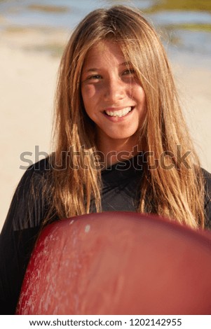 Close up shot of talented young female surfboarder likes windsurfing, carries board, has long straight hair and toothy smile, enjoys spiritual connection with nature, involved in competitive sport