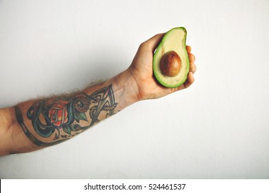 Close up shot of a tattooed man's arm with a half of ripe fresh avocado isolated on white - Powered by Shutterstock