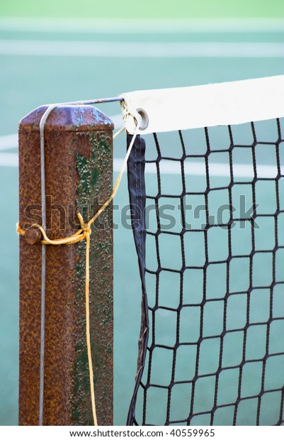 Close shot of tennis court net attached to\
rusting metal post with yellow and grey ropes.  Tennis court is out\
of focus in background.