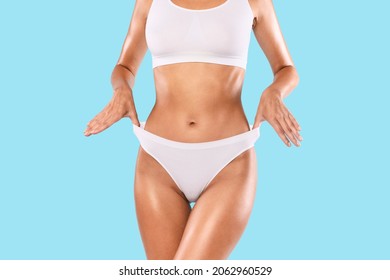 Close Up Shot Of Unrecognizable Fit Woman Wearing Lingerie Pulling Up White Panties Standing Isolated On Blue Studio Background. Torso Of Slim Female With Flat Belly In Underwear Top Bra